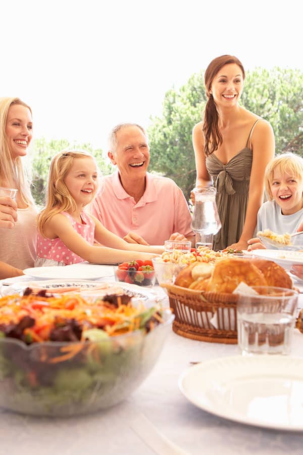 family around an outdoor patio table with a large salad and fresh rolls, drinks on display, trees in the background