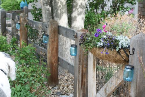 mason jars with black handles and hooks hanging from a wooden fence with a flower box holding pink and purple flowers and trees in the background