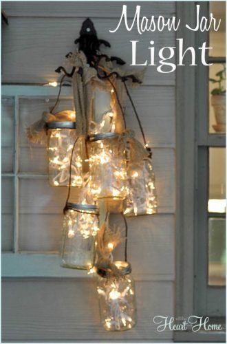 front door arrangement of mason jars with string lights inside hanging from a black iron hook on white siding