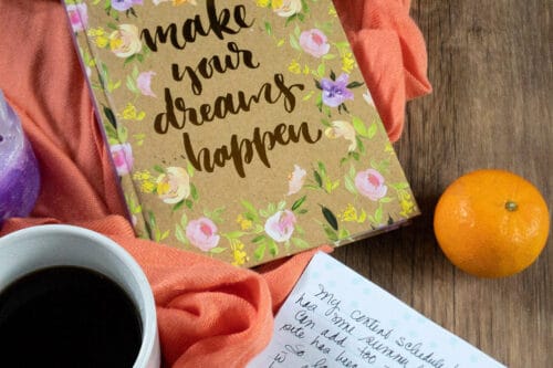 coffee, orange shawl, gold lettering on a floral patterned journal that says make your dreams happen a mandarin orange and the corner of a journal page with writing on it