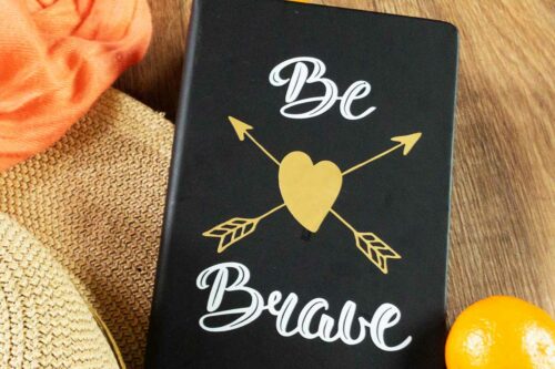 black covered journal with white and gold lettering that says be brave with a gold heart in the centre and arrows criss crossed coral shawl, sunhat and mandarin orange in the background