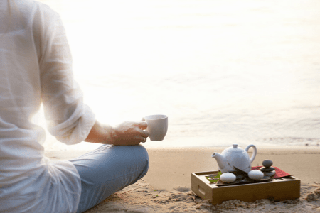 woman with tea up in hand sitting crosslegged on a beach with tea set beside her.