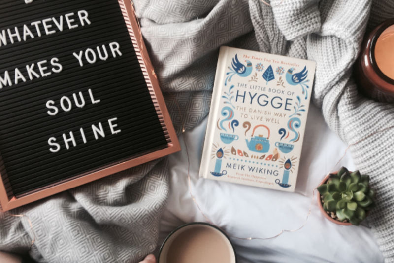 letter board that reads whatever makes your soul shine beside The Little Book of Hygge on a blanket with a candle and succulent plant to the side and bottom right 