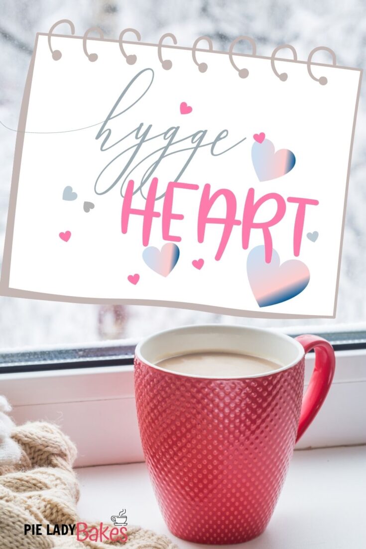 hygge heart design on a note card with a red mug of hot chocolate and blanket by window