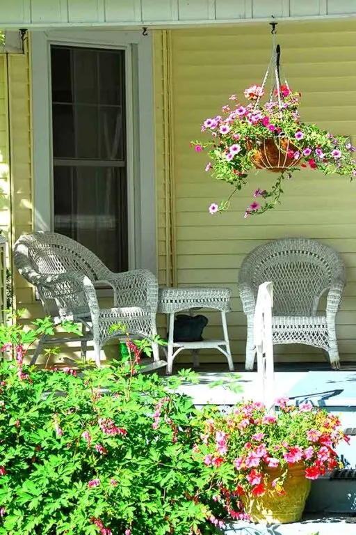 porch with white wicker furniture, a hanging plant with purple flours, yellow siding on the house, white screen door and green floral bush and more potted plants in the foreground 